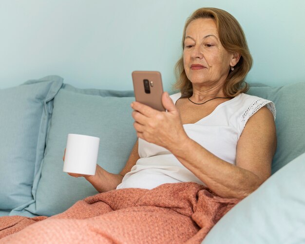 Femme âgée à la maison pendant la pandémie en dégustant une tasse de café et à l'aide de smartphone