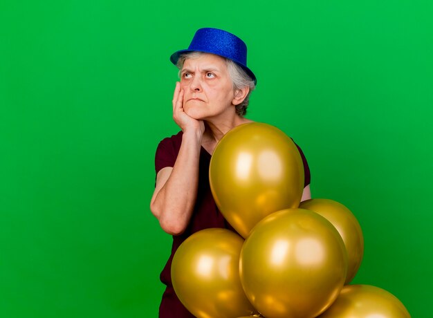 Femme âgée mécontente portant chapeau de fête met la main sur le menton debout avec des ballons d'hélium et regardant de côté sur le vert