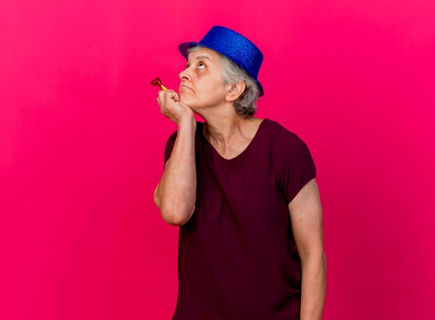 Femme âgée réfléchie portant chapeau de fête met la main sur le menton tenant le sifflet à la recherche sur rose