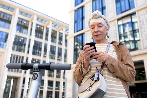 Femme âgée avec un scooter électrique dans la ville à l'aide d'un smartphone