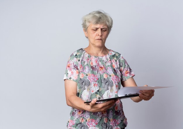 Femme âgée sérieuse tient et regarde le presse-papiers isolé sur mur blanc