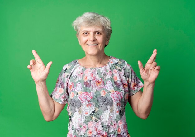 Femme âgée souriante croise les doigts isolés sur le mur vert