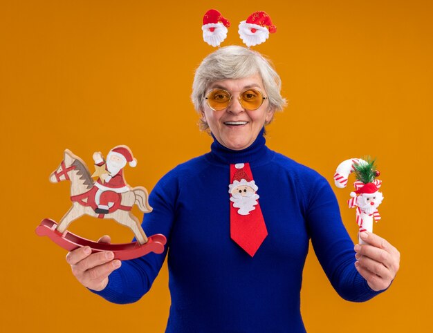 Femme âgée souriante dans des lunettes de soleil avec bandeau de père Noël et cravate de père Noël tenant le père Noël sur la décoration de cheval à bascule et canne en bonbon isolé sur fond orange avec espace copie