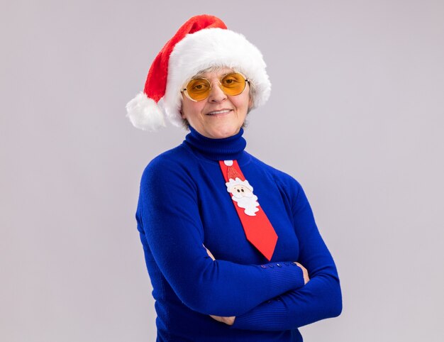 Femme âgée souriante dans des lunettes de soleil avec Bonnet de Noel et cravate de Père Noël debout avec les bras croisés isolé sur fond blanc avec espace copie