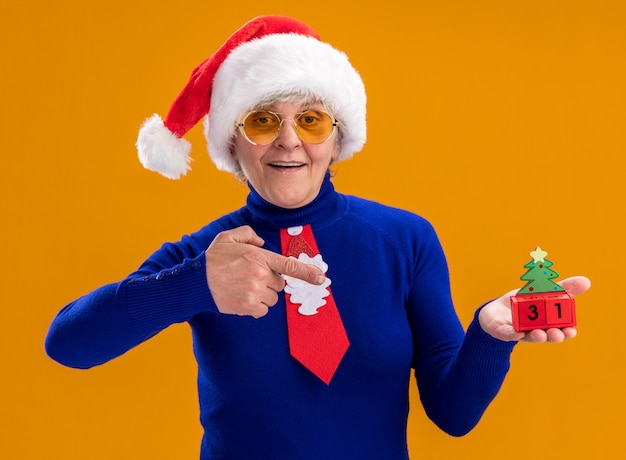 Femme âgée souriante dans des lunettes de soleil avec Bonnet de Noel et cravate de Père Noël tenant et pointant sur ornement d'arbre de Noël isolé sur fond orange avec espace copie