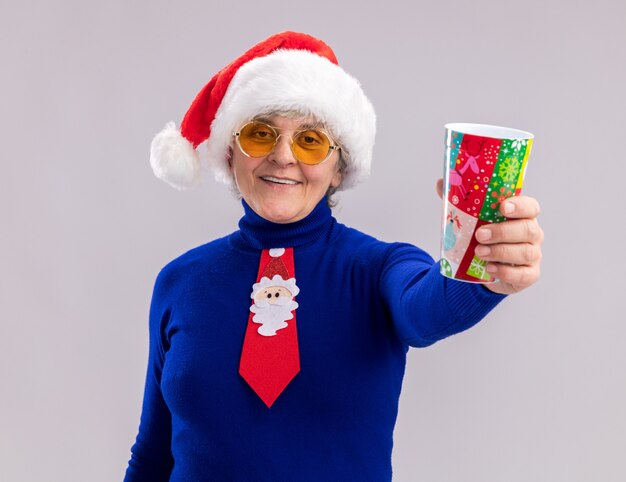 Femme âgée souriante dans des lunettes de soleil avec Bonnet de Noel et cravate de Père Noël tenant une tasse de papier