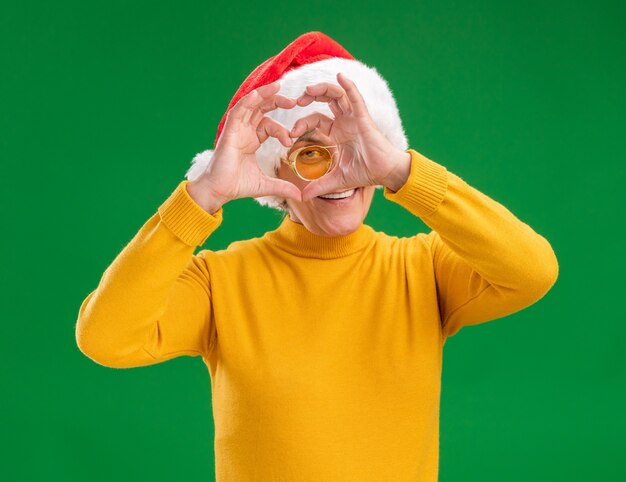 Femme âgée souriante dans des lunettes de soleil avec bonnet de Noel par signe de coeur