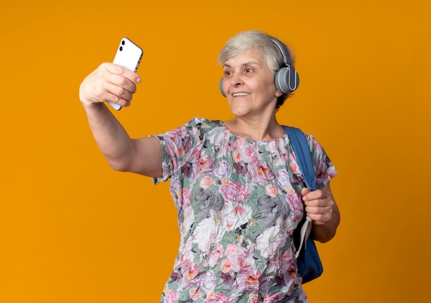 Femme âgée Souriante Sur Des écouteurs Portant Sac à Dos Tient Et Regarde Le Téléphone Prenant Selfie Isolé Sur Mur Orange