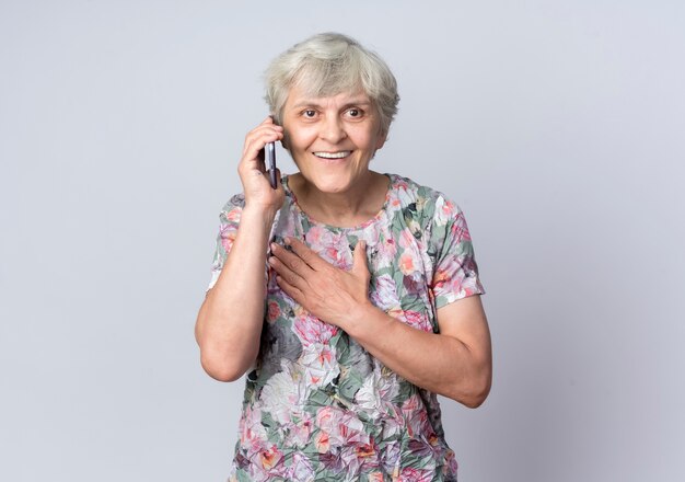 Femme âgée souriante met la main sur le menton en parlant au téléphone isolé sur un mur blanc