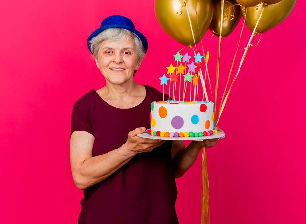 Femme âgée souriante portant chapeau de fête détient des ballons d'hélium et un gâteau d'anniversaire regardant la caméra sur rose