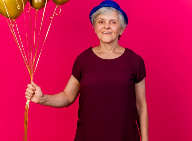 Femme âgée souriante portant chapeau de fête détient des ballons d'hélium sur rose