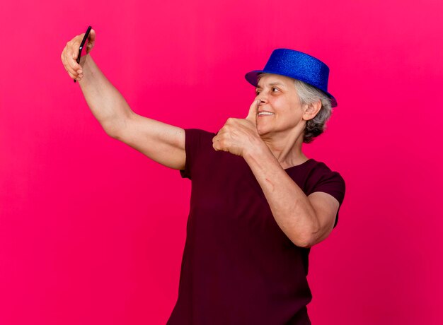 Femme âgée souriante portant chapeau de fête pouces vers le haut tenant et regardant le téléphone sur rose