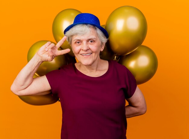 Femme âgée souriante portant des gestes de chapeau de fête signe de la main de la victoire et détient des ballons d'hélium