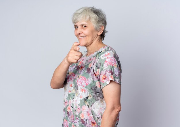 Femme âgée souriante se tient sur le côté pointant isolé sur un mur blanc