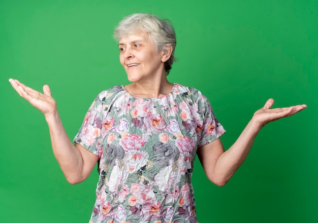 Femme âgée souriante se tient avec les mains levées en regardant côté isolé sur mur vert