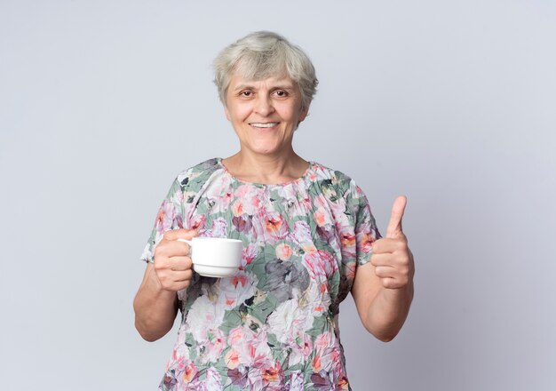 Femme âgée souriante tient la tasse et les pouces vers le haut isolé sur un mur blanc