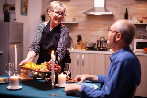 Femme âgée tenant une assiette en bois avec et regardant son mari pendant le dîner de fête. Vieux couple de personnes âgées parlant, assis à table dans la cuisine, savourant le repas, célébrant leur anniversaire.