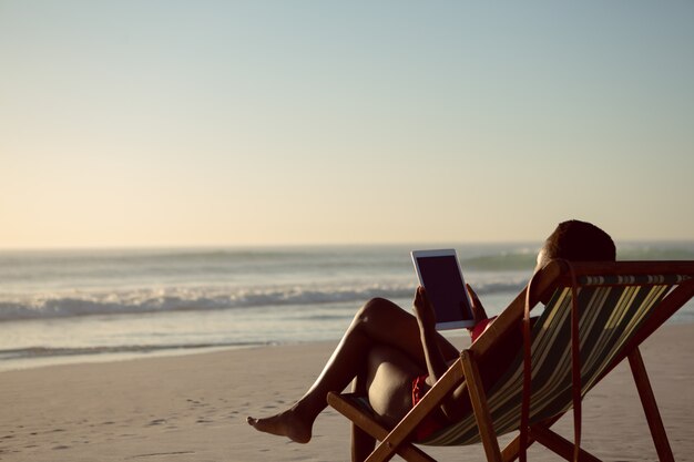 Femme à l'aide de tablette numérique tout en vous relaxant dans une chaise de plage sur la plage
