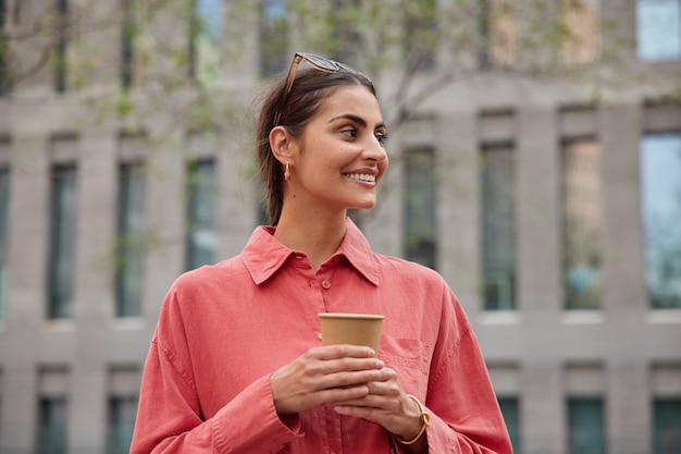 une femme aime le chocolat chaud aromatique dans une tasse à emporter se promène dans la ville regarde ailleurs avec une expression heureuse porte des poses de chemise