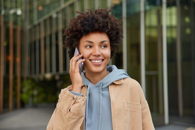 La femme aime la communication cellulaire sourit positivement habillée pose avec désinvolture à l'extérieur fait un appel téléphonique