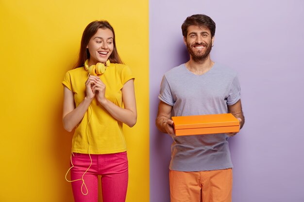 Une femme à l'air agréable garde les mains en prévision, regarde curieusement la boîte en carton, étant intéressante ce qui est à l'intérieur. Heureux jeune homme tient peu de contrainer ou paquet, donne cadeau à petite amie