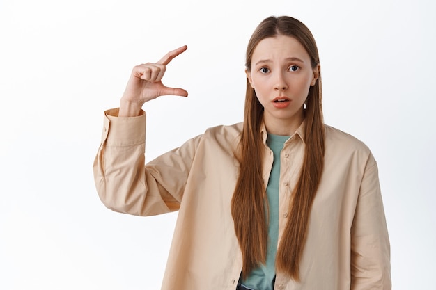 La femme a l'air confuse alors qu'elle montre un tout petit geste, une petite chose sur le fond, regarde devant perplexe, déçue par la taille, debout sur un mur blanc