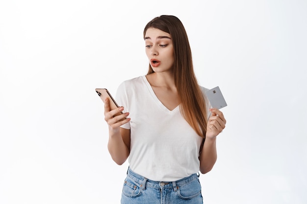 Une femme a l'air surprise de son smartphone tout en payant avec une carte de crédit, en faisant des achats en ligne, en haletant et en haussant les sourcils, intriguée par quelque chose sur l'écran du mobile, debout sur un mur blanc