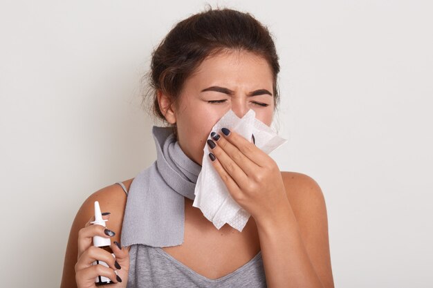 femme allergique malade se moucher, avoir la grippe ou attraper froid, éternuer dans un mouchoir, posant les yeux fermés isolé sur blanc, tenant un spray nasal à la main.