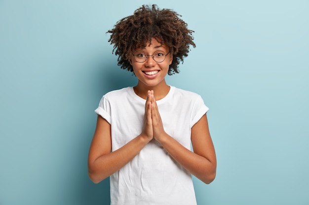 Une femme amicale à la peau sombre tient les paumes ensemble sur la poitrine, montre le geste de namaste, demande de l'aide, a une expression heureuse, porte un t-shirt blanc, des lunettes optiques, isolées sur un mur bleu.