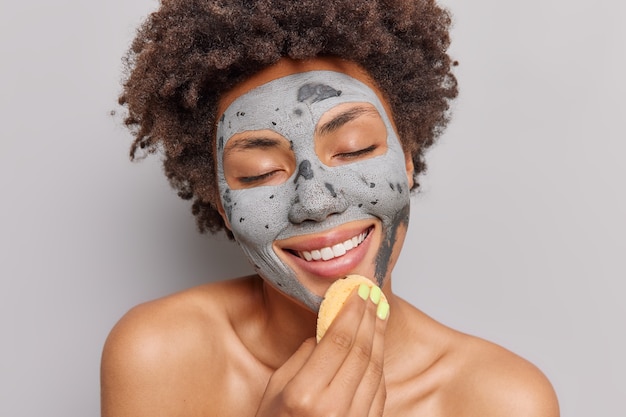 Photo gratuite la femme applique un masque d'argile avec des sourires d'éponge cosmétique apprécie doucement les procédures de soins de la peau ferme les yeux du plaisir pose nue à l'intérieur. notion de beauté.