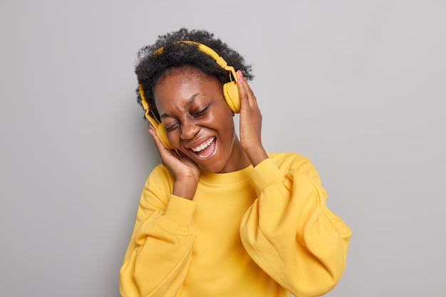 Photo gratuite la femme apprécie le son stéréo dans des écouteurs modernes sans fil porte un pull jaune décontracté garde les yeux fermés aime écouter de la musique isolée sur fond gris