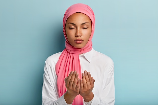 Une femme arabe fidèle à la peau foncée garde les mains dans un geste de prière, demande à Allah une bonne santé, croit au bien-être a la tête voilée, porte une chemise blanche garde les yeux fermés bénéficie d'une atmosphère paisible