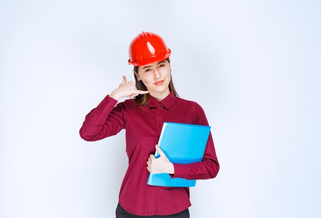 Femme architecte en casque rouge tenant un dossier bleu et posant devant la caméra.