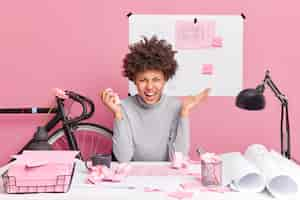 Photo gratuite une femme architecte en colère pose à table avec des plans de papiers et des restes en colère de trouver une erreur dans son travail de projet s'exclame avec des poses d'expressio outrées contre le mur rose dans l'espace de coworking