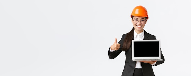 Femme architecte professionnelle souriante responsable de la construction à l'usine montrant un diagramme positif graphique faire le pouce levé en approbation et tenant un ordinateur portable face à l'écran porter un casque de sécurité