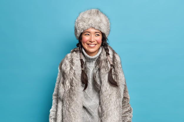 Femme arctique positive avec deux nattes peignées sourit heureusement a la bonne humeur vêtue de vêtements d'hiver chauds bénéficie de l'heure d'hiver et marche en plein air pendant une journée glaciale isolée sur mur bleu