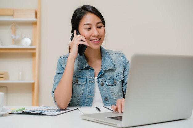 Femme asiatique belle entreprise intelligente en vêtements décontractés travaillant sur ordinateur portable et parler au téléphone tout en étant assis sur la table dans le bureau créatif.