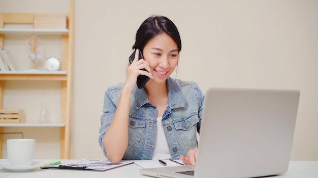 Femme asiatique belle entreprise intelligente en vêtements décontractés travaillant sur ordinateur portable et parler au téléphone tout en étant assis sur la table dans le bureau créatif.