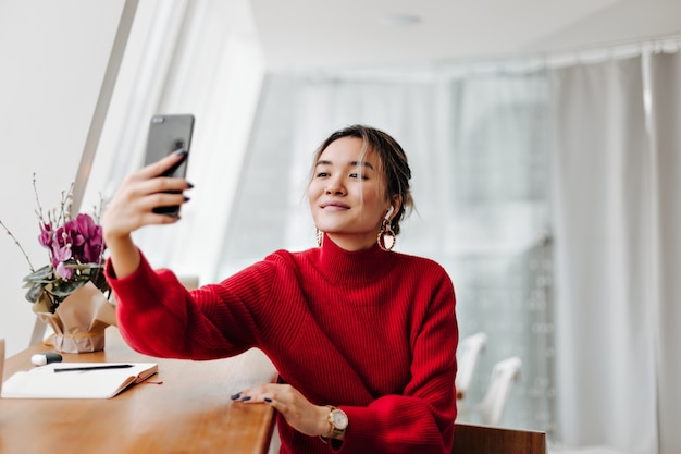 Femme asiatique blonde en boucles d'oreilles élégantes et pull rouge fait selfie contre fenêtre