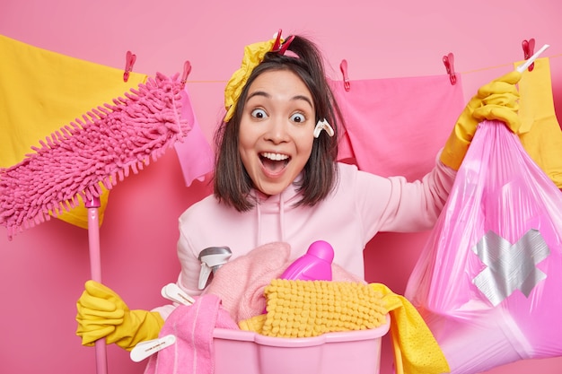 Une femme asiatique brune et gaie, surprise, garde la bouche ouverte, porte un sac poubelle et pose une vadrouille près d'un panier à linge près d'une corde à linge contre un mur rose