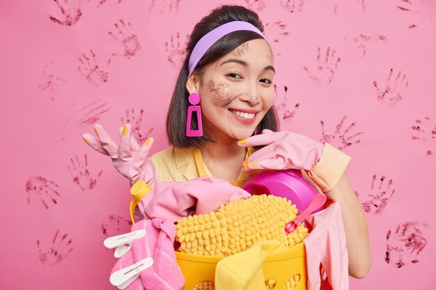 Une femme asiatique brune heureuse se penche sur le panier à linge sourit joyeusement d'être sale après avoir fait le ménage porte des gants de protection en caoutchouc bandeau isolé sur un mur rose