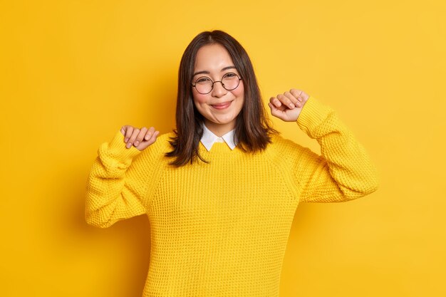 Une femme asiatique brune heureuse se sent soulagée, lève les bras et sourit de plaisir porte des lunettes rondes.