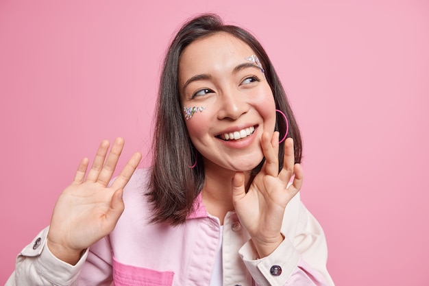 Une femme asiatique brune positive sourit largement a un visage de dents blanches parfaites orné de pierres brillantes, une expression joyeuse et rêveuse garde les mains levées porte une veste en jean isolée sur un mur rose