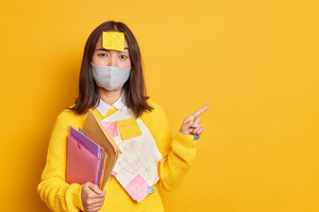 Une femme asiatique brune sérieuse a un autocollant sur la tête tente d'apprendre des informations pour réussir l'examen porte un masque jetable pour éviter les points de coronavirus dans un espace vide vide