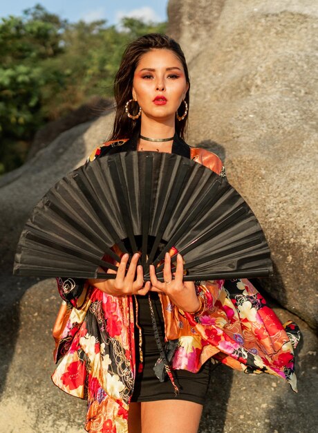 Femme asiatique en chemisier kimono en soie colorée tenant un ventilateur noir et posant sur la plage avec des rochers