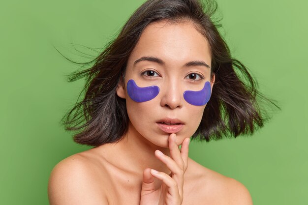 Une femme asiatique a les cheveux noirs flottant dans l'air applique des patchs d'hydrogel bleu sous les yeux subit des soins de la peau