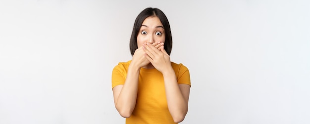 Une femme asiatique choquée couvre la bouche avec les mains semblant surprise par l'expression du visage sans voix debout en t-shirt jaune sur fond blanc