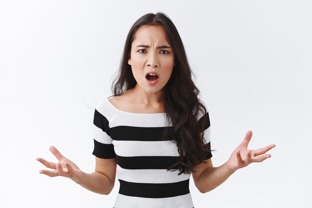 Une femme asiatique en colère énervée et énervée en t-shirt rayé a un énorme regard de plainte agacé et dérangé fronçant les mains écartées sur le côté haletant offensé et frustré debout fond blanc