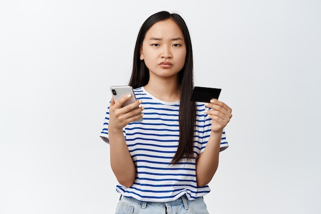 Femme asiatique en colère tenant une carte de crédit et un téléphone portable fronçant les sourcils déçu debout sur fond blanc