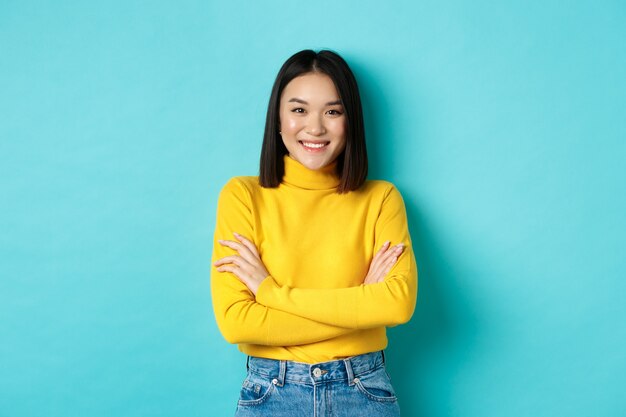 Une femme asiatique confiante et élégante croise les bras sur la poitrine et souriante, debout sur fond bleu.